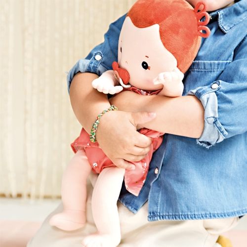 Detail van een kind dat de Lilliputiens lappenpop Rose met rode jurk en speen vasthoudt.