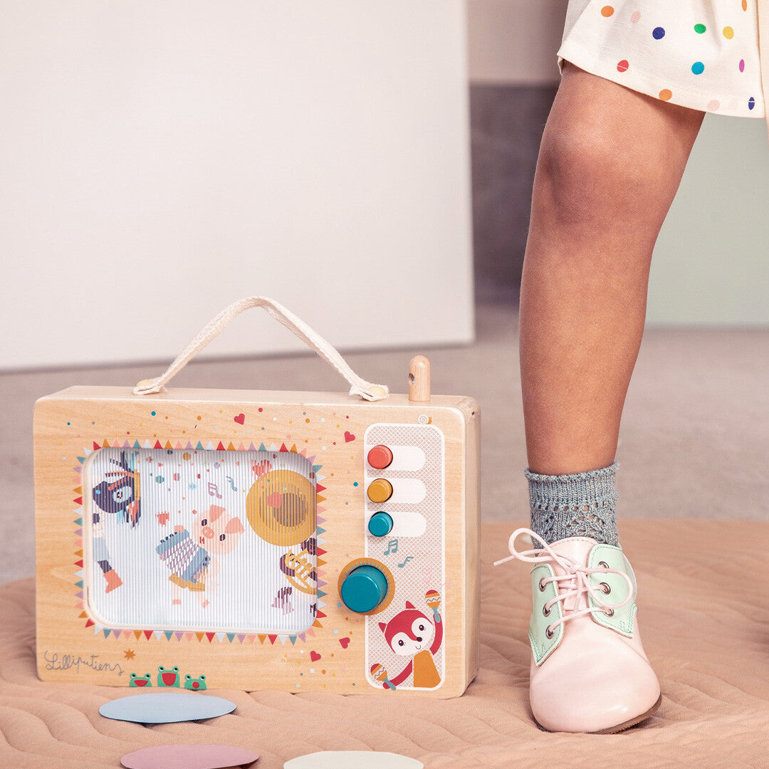 Lilliputiens houten muziekdoos staat op een mat naast de voet van een kind met een roze schoen.