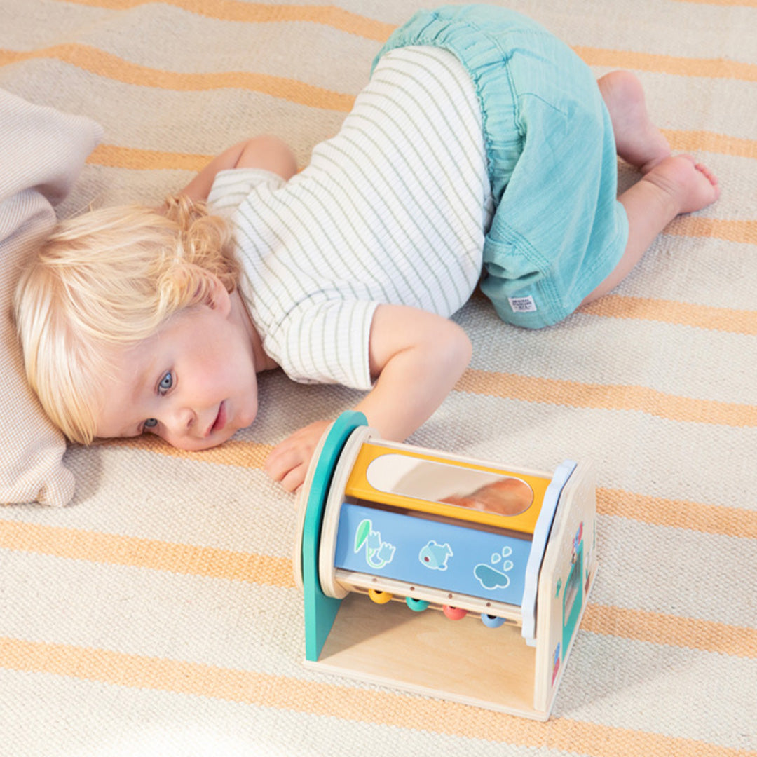 Een baby ligt op de grond en kijkt naar de Lilliputiens houten activiteiten- kleurenroller met blauwe figuren.
