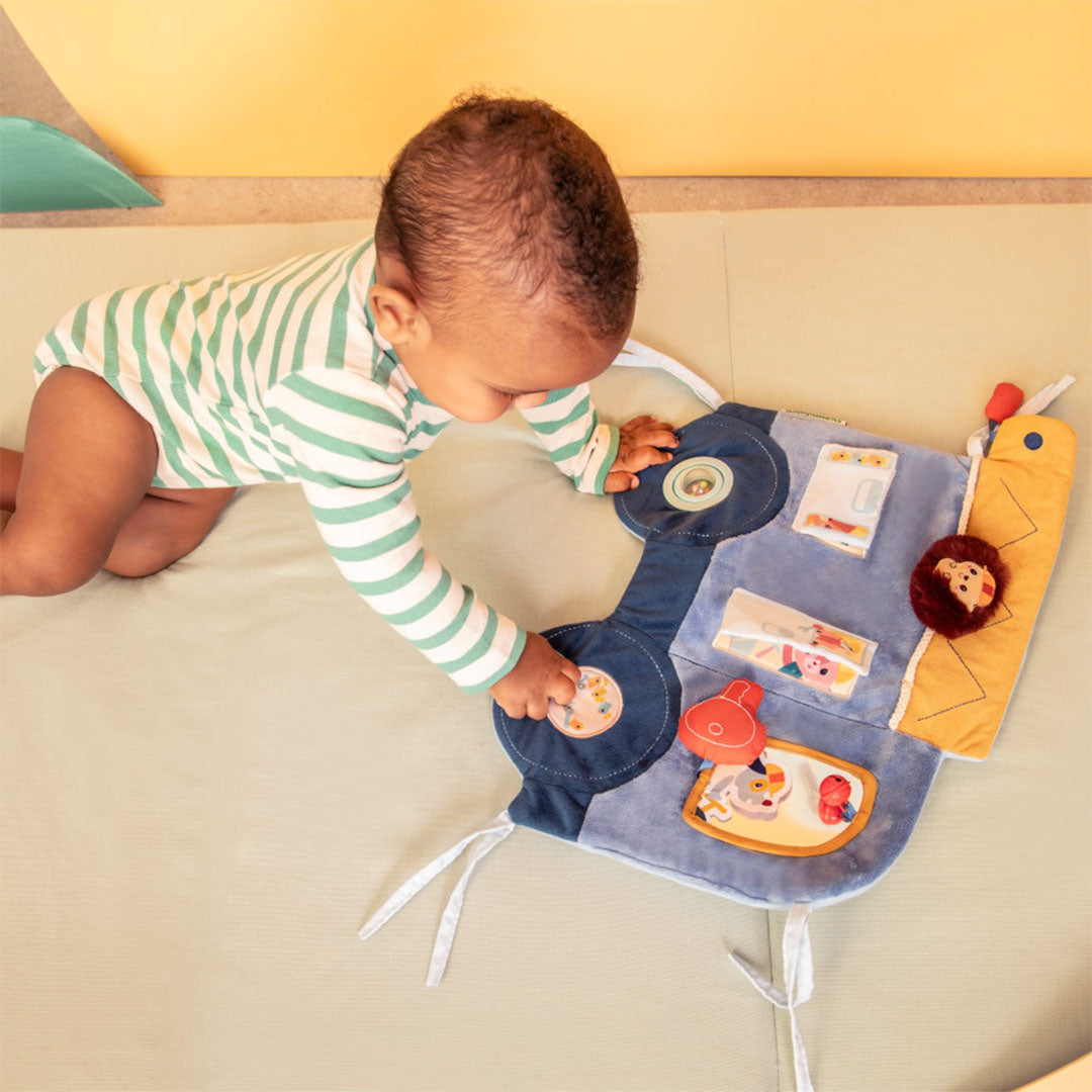 Een baby op de buik reikt naar het Lilliputiens activiteitenpaneel Jules de klusjesman dat op een lichtgroene ondergrond ligt.