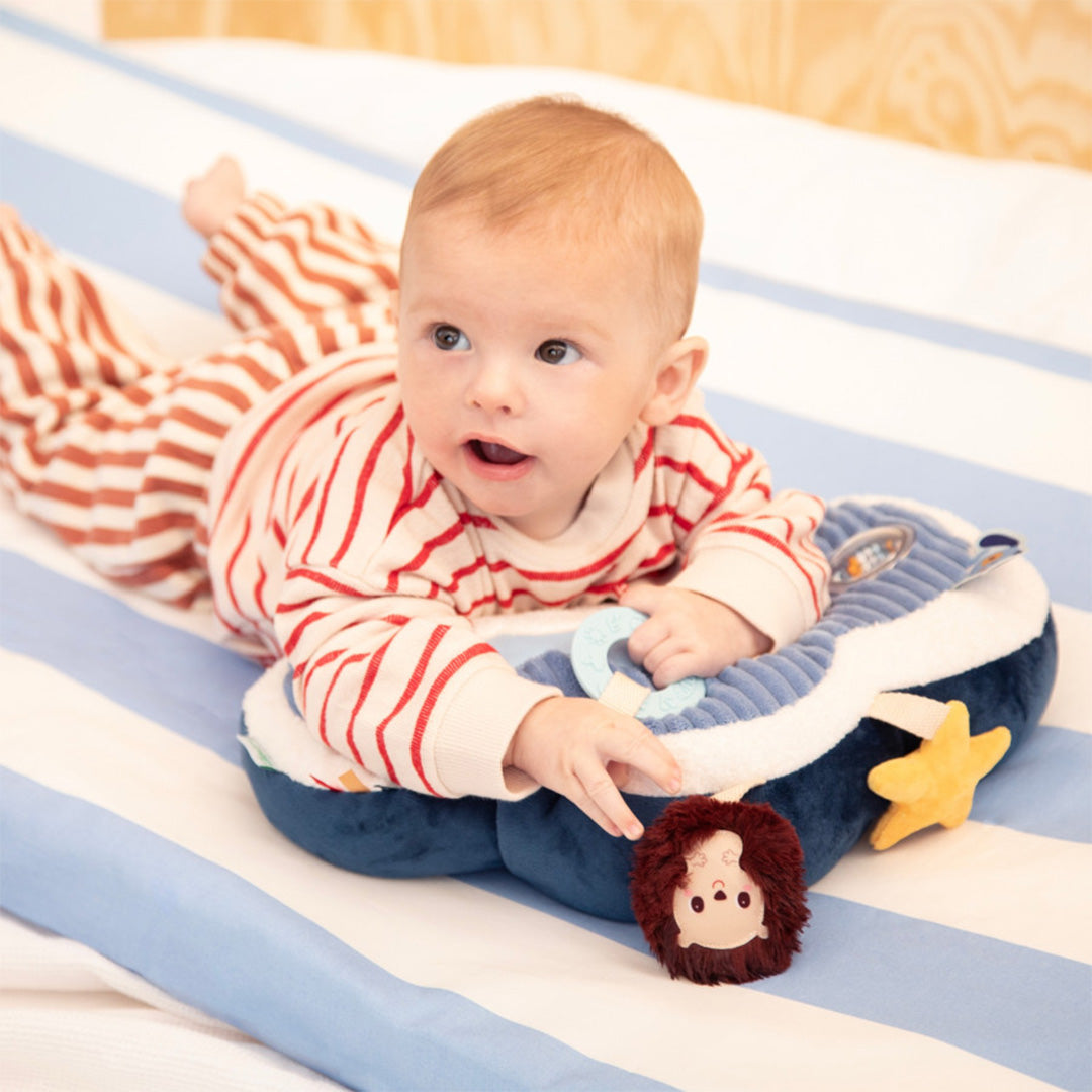 Een baby speelt op de buik met het Lilliputiens activiteitenkussen Jules de hond, met de hand bij de bijtring.