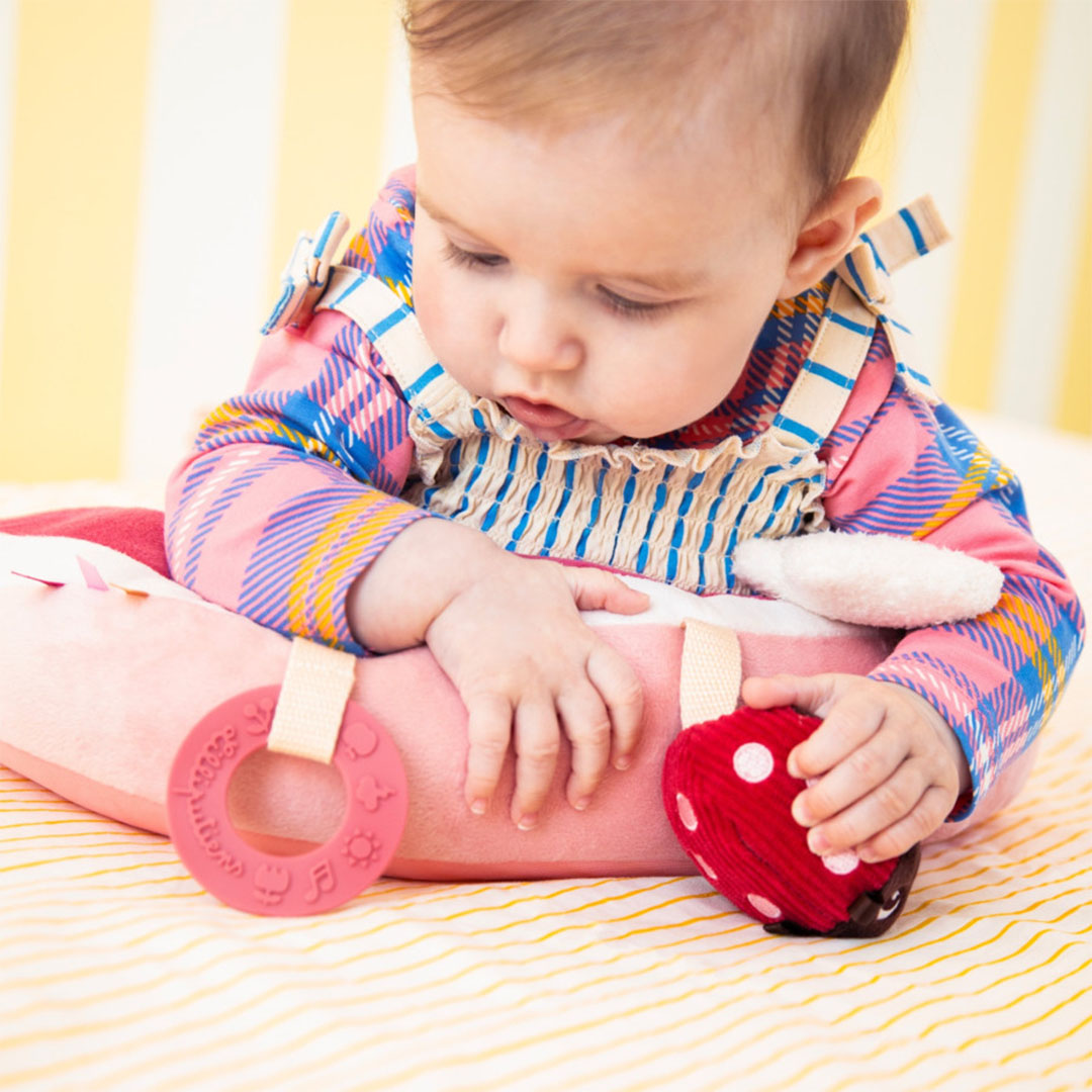 Een baby speelt met het Lilliputiens activiteitenkussen, vasthoudend aan het lieveheersbeestje en een roze bijtring.