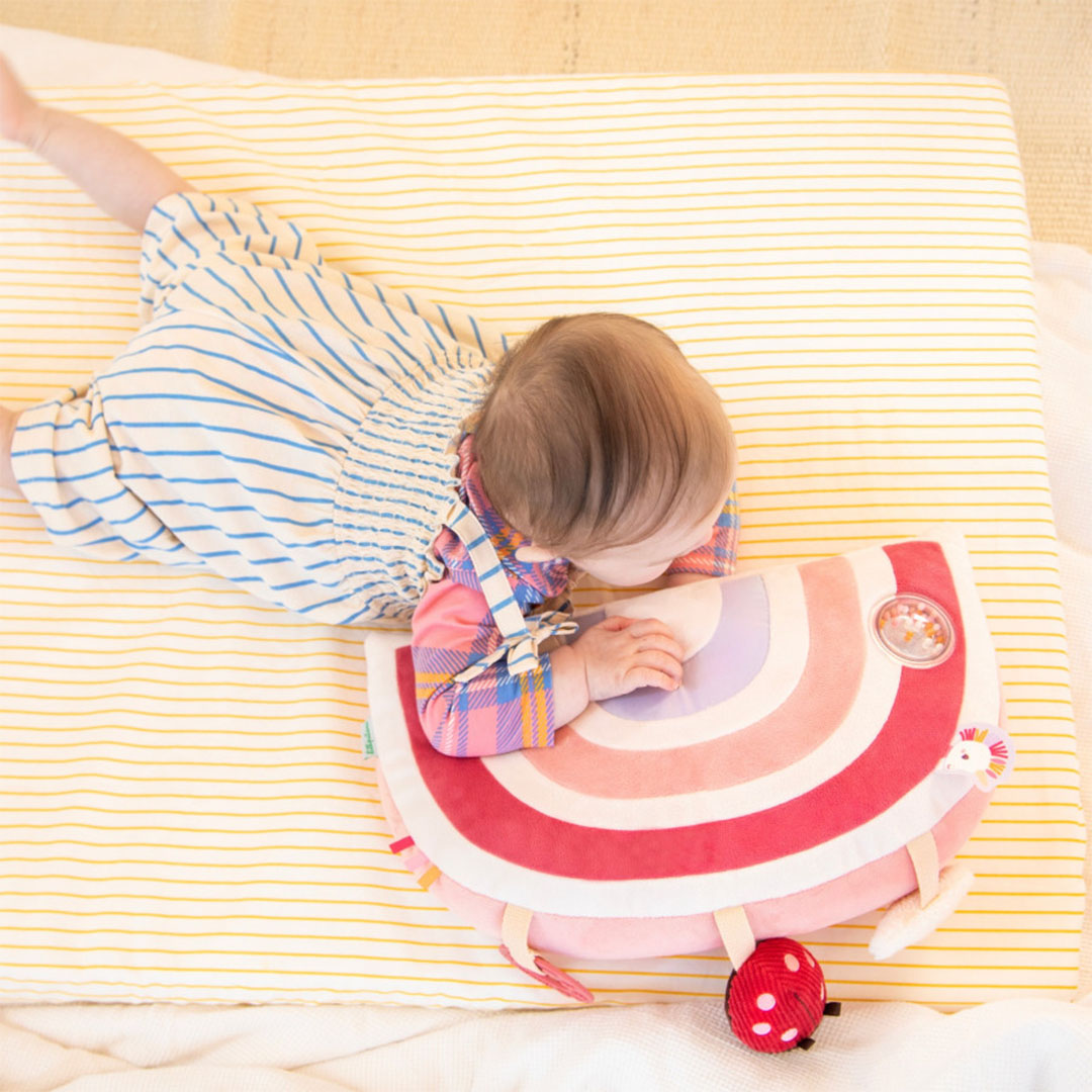 Een baby ligt op de buik en speelt met het Lilliputiens activiteitenkussen in regenboogvorm.