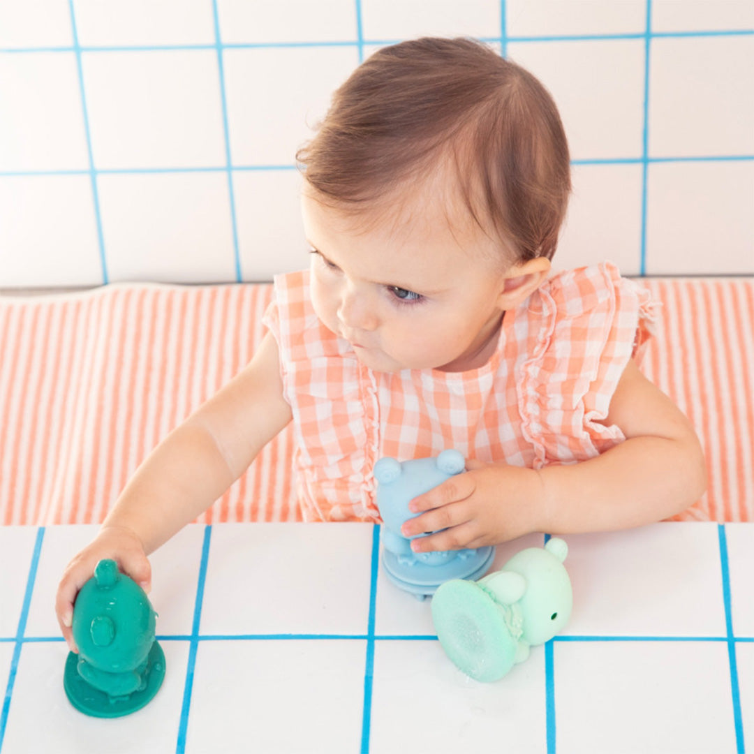 Een baby speelt in de badkamer met twee Lilliputiens badkikkers, een groene en een lichtblauwe.