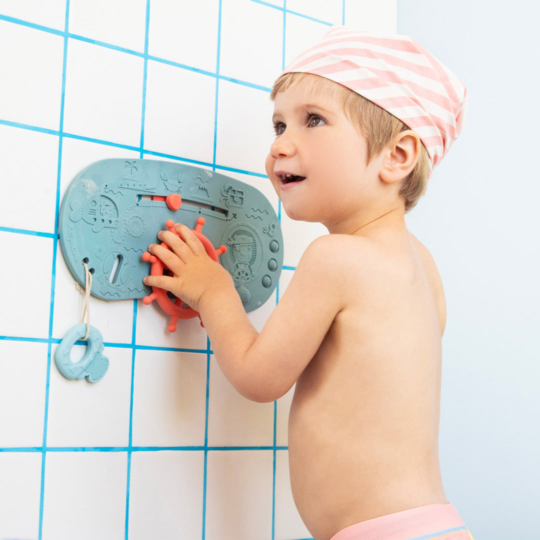 Een kind speelt met het Lilliputiens interactieve badspeelgoed, een blauw piratenpaneel, bevestigd aan een tegelmuur.