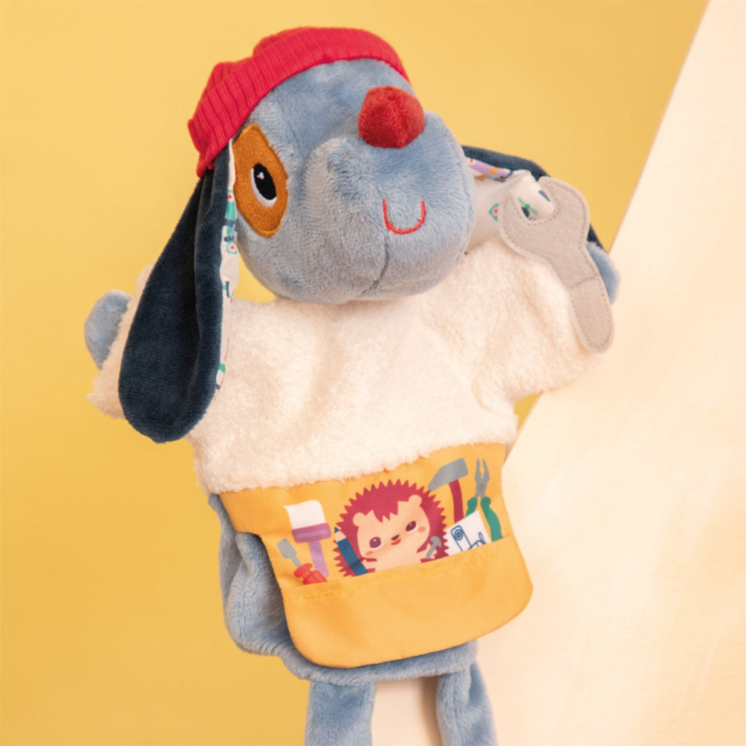 Lilliputiens handpop Jules, een blauwe hond met rode muts en gele gereedschapsapron, schuin gefotografeerd op een gele achtergrond.