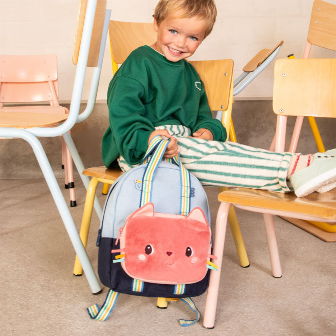 Een jongen zit op een stoel en houdt een blauwe rugzak vast met het roze lilliputiens kindertasje met kattengezicht eraan bevestigd.