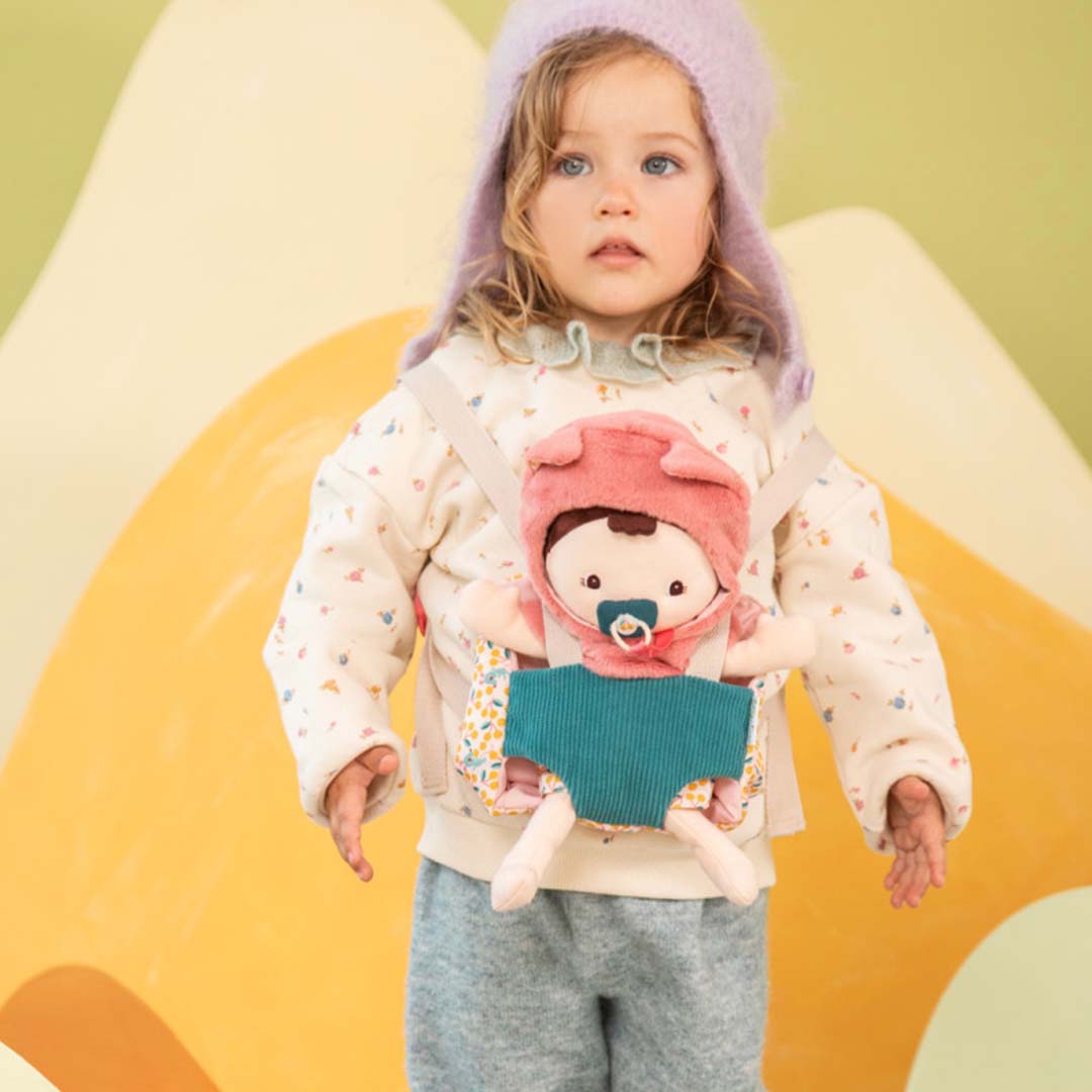 Een kind draagt de Lilliputiens lappenpop Jeanne in de gebloemde draagzak, staand voor een kleurrijke achtergrond.