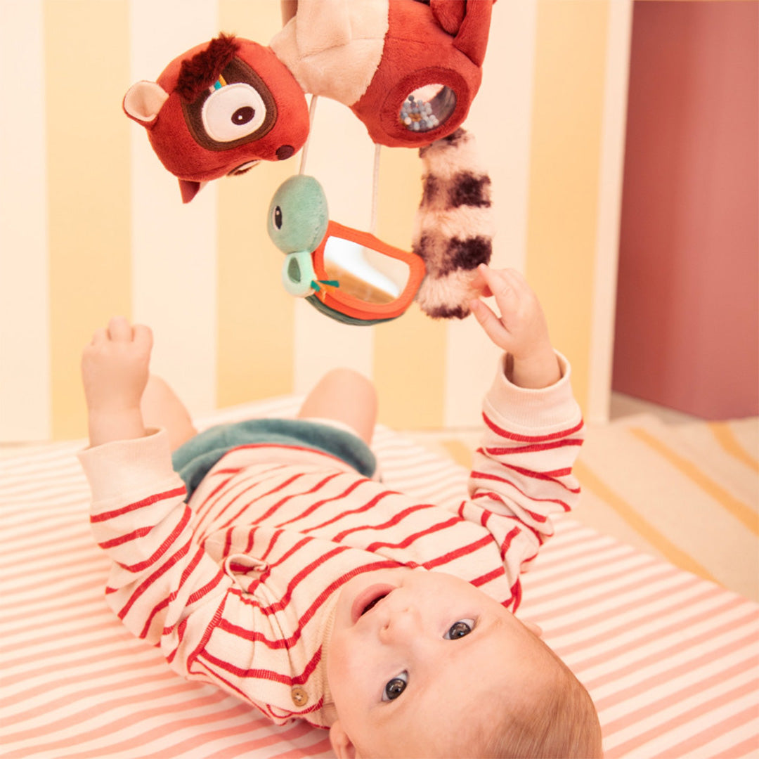 Een baby reikt liggend naar de hangende activiteitenhanger Elvis de maki.