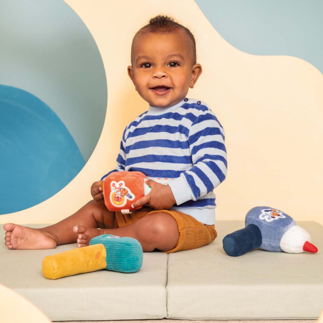 Een lachende baby zit op de grond met een oranje Lilliputiens speelgoedgereedschap en de hamer rammelaar ernaast.