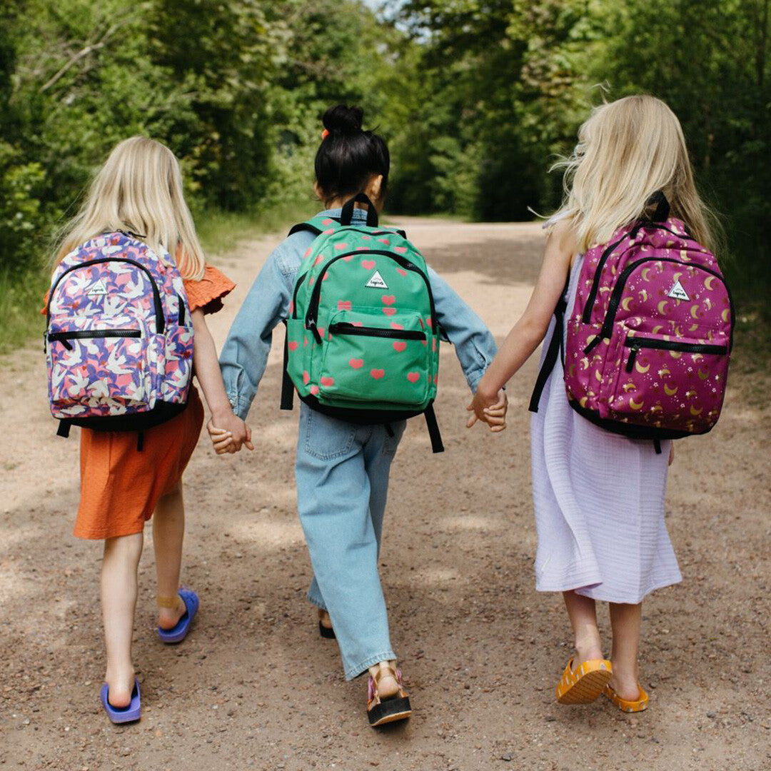 Drie kinderen lopen hand in hand met verschillende little legends kinderrugzakken op de rug, waaronder de groene met roze harten.