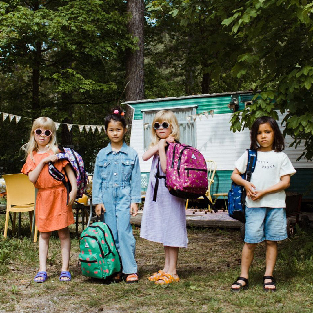 Vier kinderen staan buiten met verschillende little legends kinderrugzakken, waaronder de groene met roze harten op de grond.
