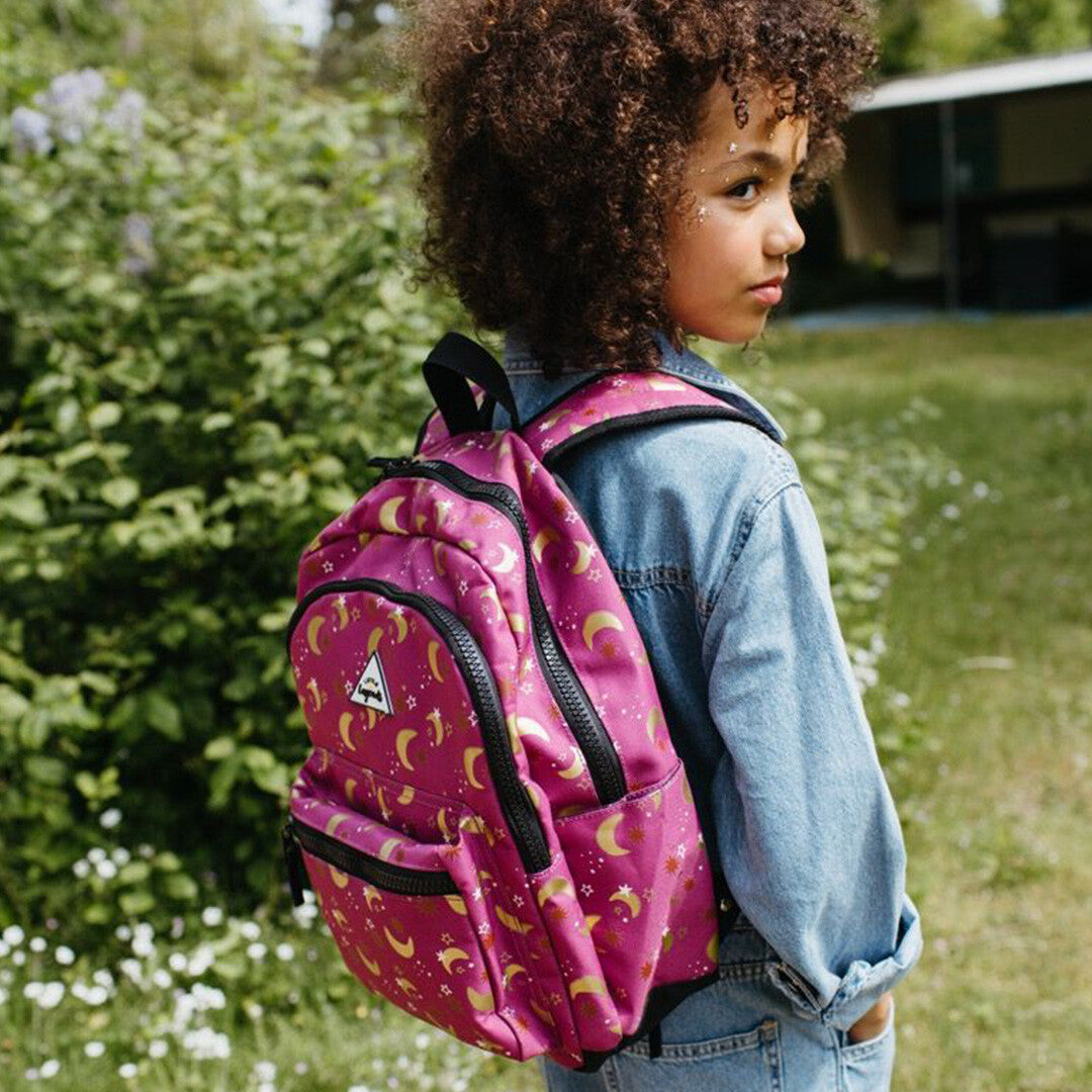 Een kind draagt de roze kinderrugzak met maan- en sterrenpatroon schuin op de rug in een groene omgeving.