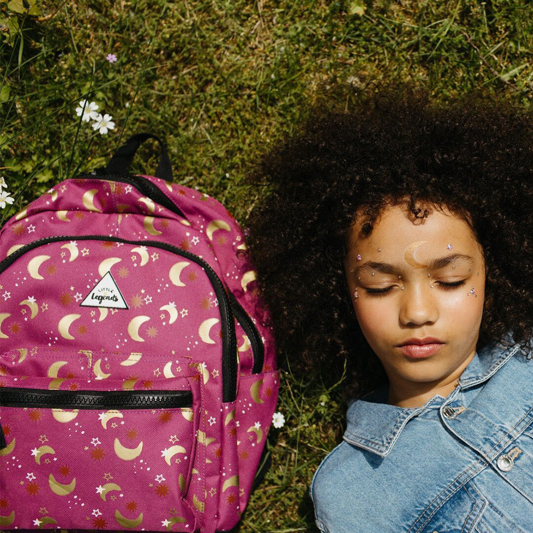 Een kind ligt op het gras naast de roze kinderrugzak met maan- en sterrenpatroon.