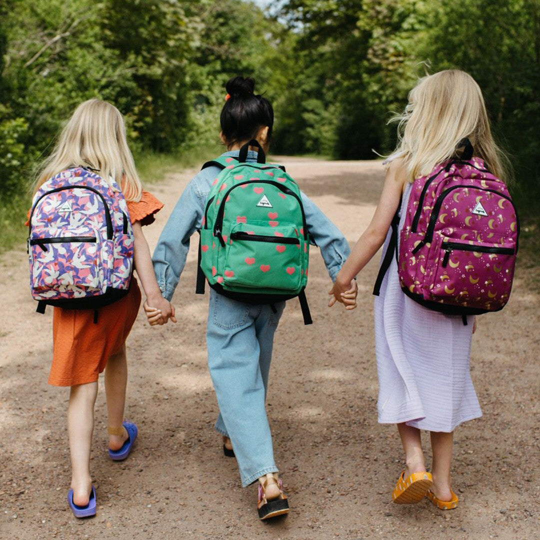 Drie kinderen lopen hand in hand over een pad met verschillende kinderrugzakken, waaronder de roze met maan- en sterrenpatroon.