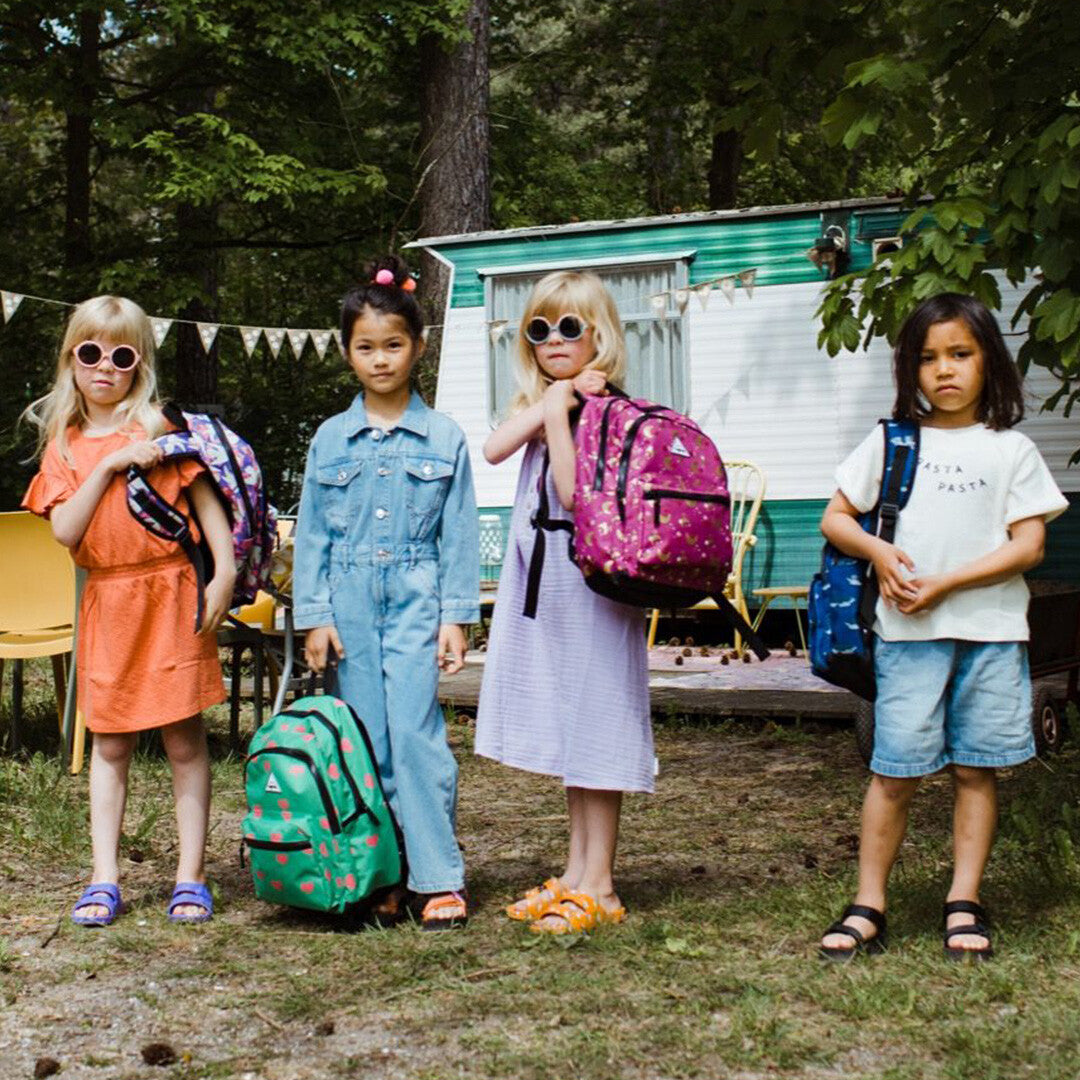 Vier kinderen staan voor een caravan met verschillende kinderrugzakken, waaronder de roze met maan- en sterrenpatroon.