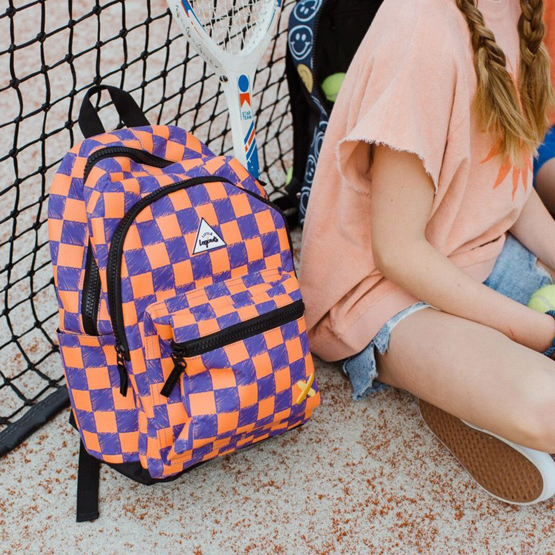 Little Legends kinderrugzak met oranje en paars checkerboard patroon staat op de grond naast een kind.