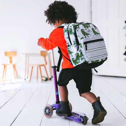 Een kind rijdt op een scooter met de Little Legends schoolrugzak met dinosaurusprint op de rug.