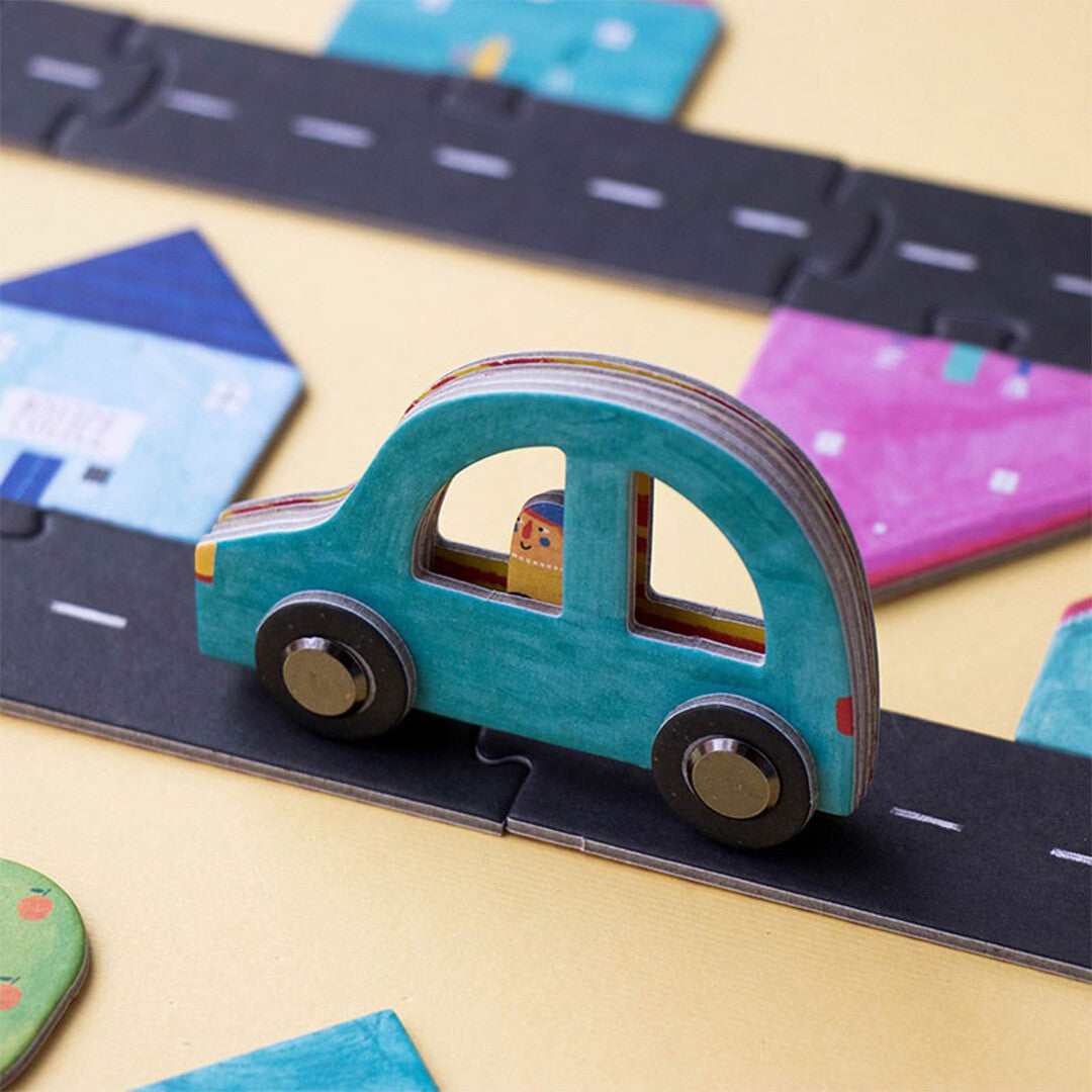 Een close-up van een blauwe houten auto op een wegdeel van de londji vloerpuzzel, met huizen op de achtergrond.