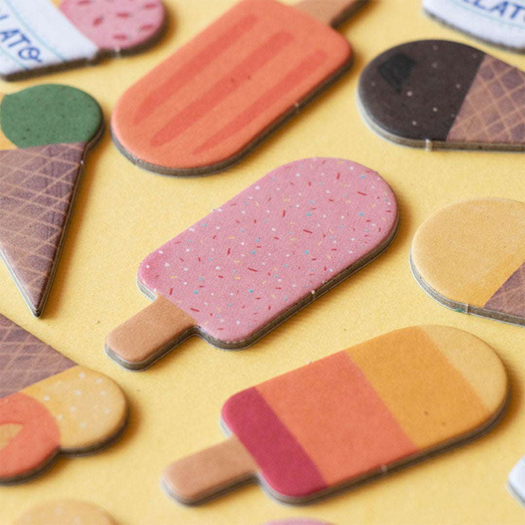 Detail van verschillende kartonnen ijsjes en waterijsjes, speelstukken van het Gelato behendigheidsspel, op een gele achtergrond.