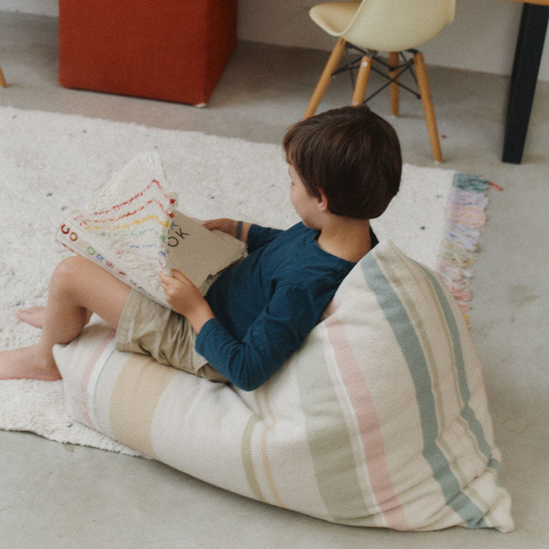 Een kind zit op de Lorena Canals poef Cleo met strepen en leest een boek in een kamer.