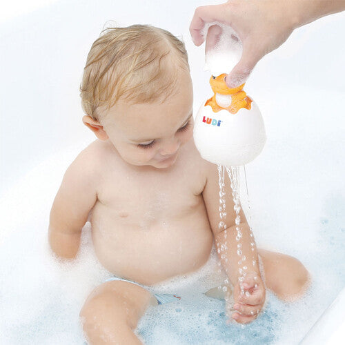 Een hand spuit water met het Ludi badspeeltje dino ei over een baby in een bad met schuim.