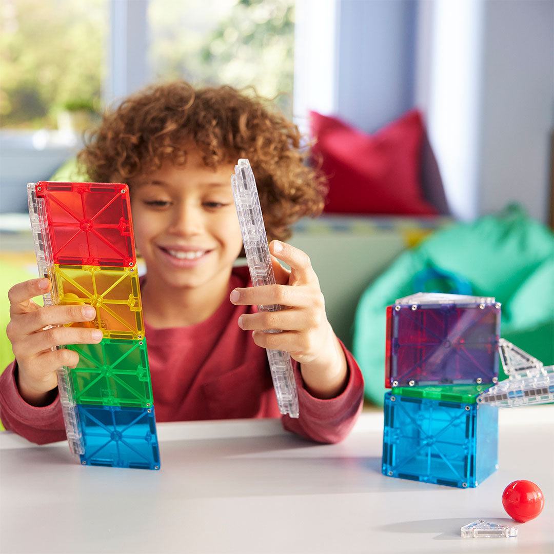 Een lachend kind bouwt met kleurrijke Magna-Tiles magnetische tegels op een tafel.