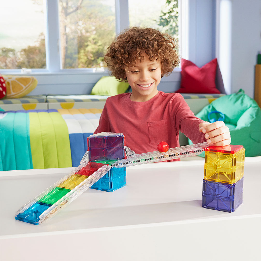 Een kind speelt met een veelkleurige Magna-Tiles knikkerbaan en laat een rode bal rollen.