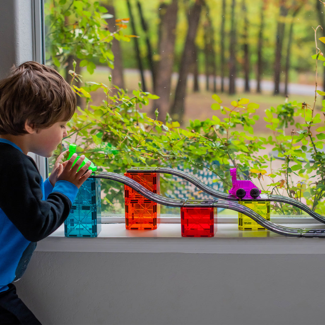 Een jongen speelt met de Magna-Tiles magnetische autobaan op een vensterbank voor een raam met uitzicht op groen.