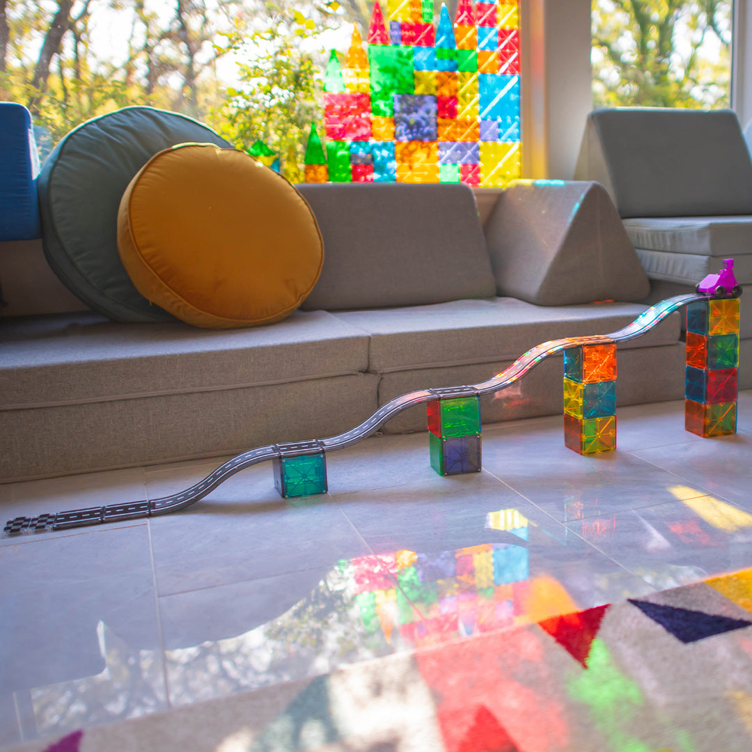 Een lange Magna-Tiles magnetische autobaan met kleurrijke ondersteuningen op de vloer, met zonlicht dat door de magnetische tegels schijnt.