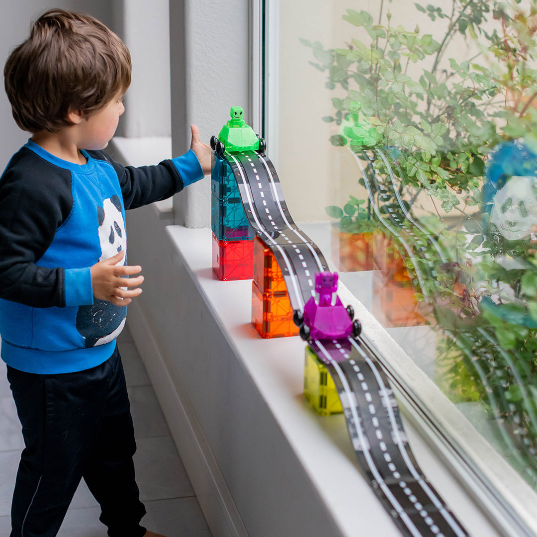 Een jongen kijkt naar de Magna-Tiles magnetische autobaan downhill set op de vensterbank, met twee auto's op de banen.