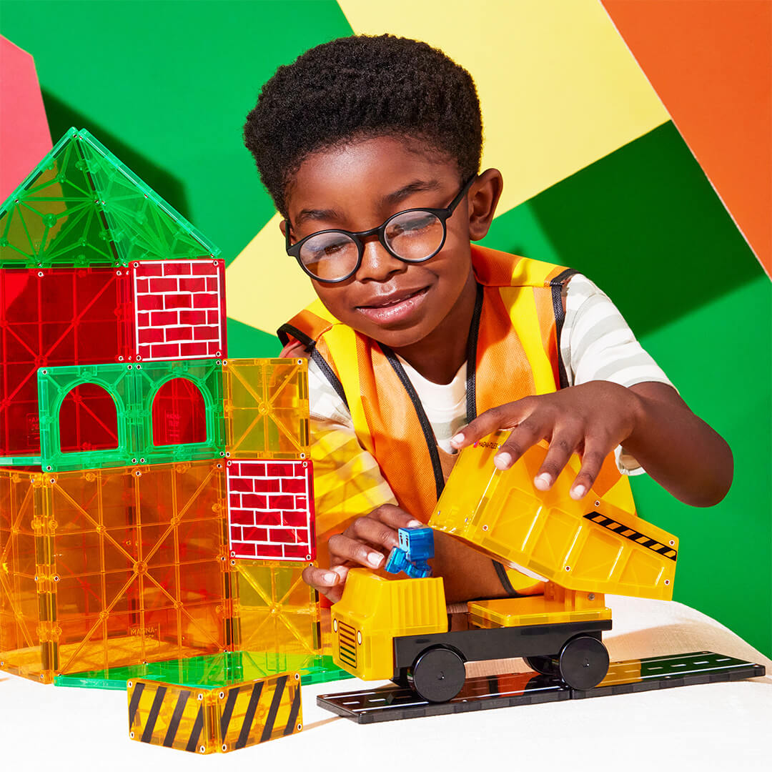 Een lachende jongen met een bouwvest speelt met de Magna-Tiles Builder XL magnetische tegels en een gele kiepwagen.