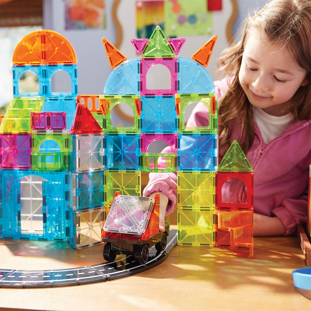 Een meisje speelt met de Magna-Tiles City Center bouwset, waarbij ze een voertuig over een weg beweegt naast een kleurrijk gebouw.