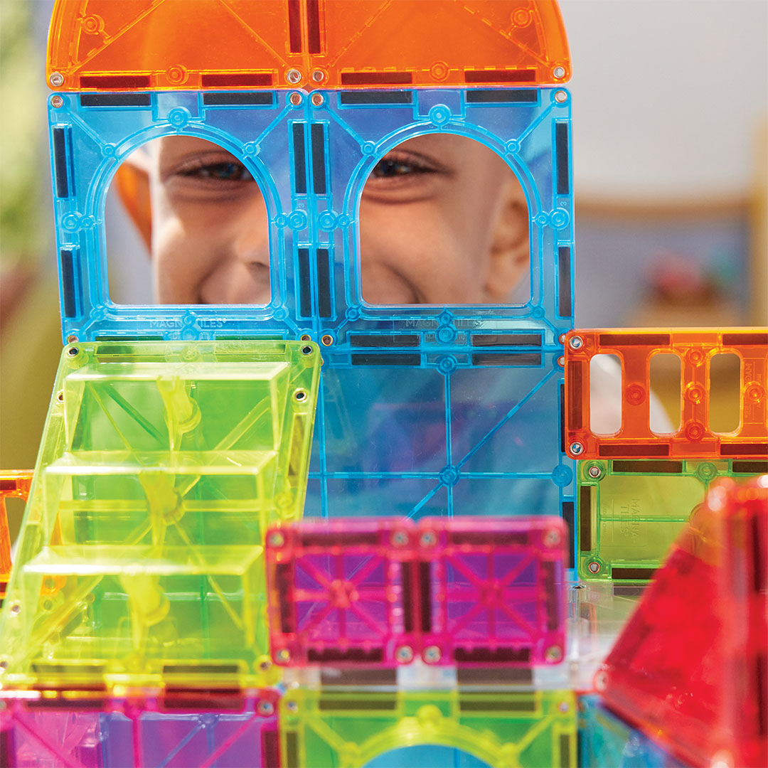 Een lachende jongen kijkt door een raam van een gebouw gemaakt met Magna-Tiles City Center magnetische tegels.