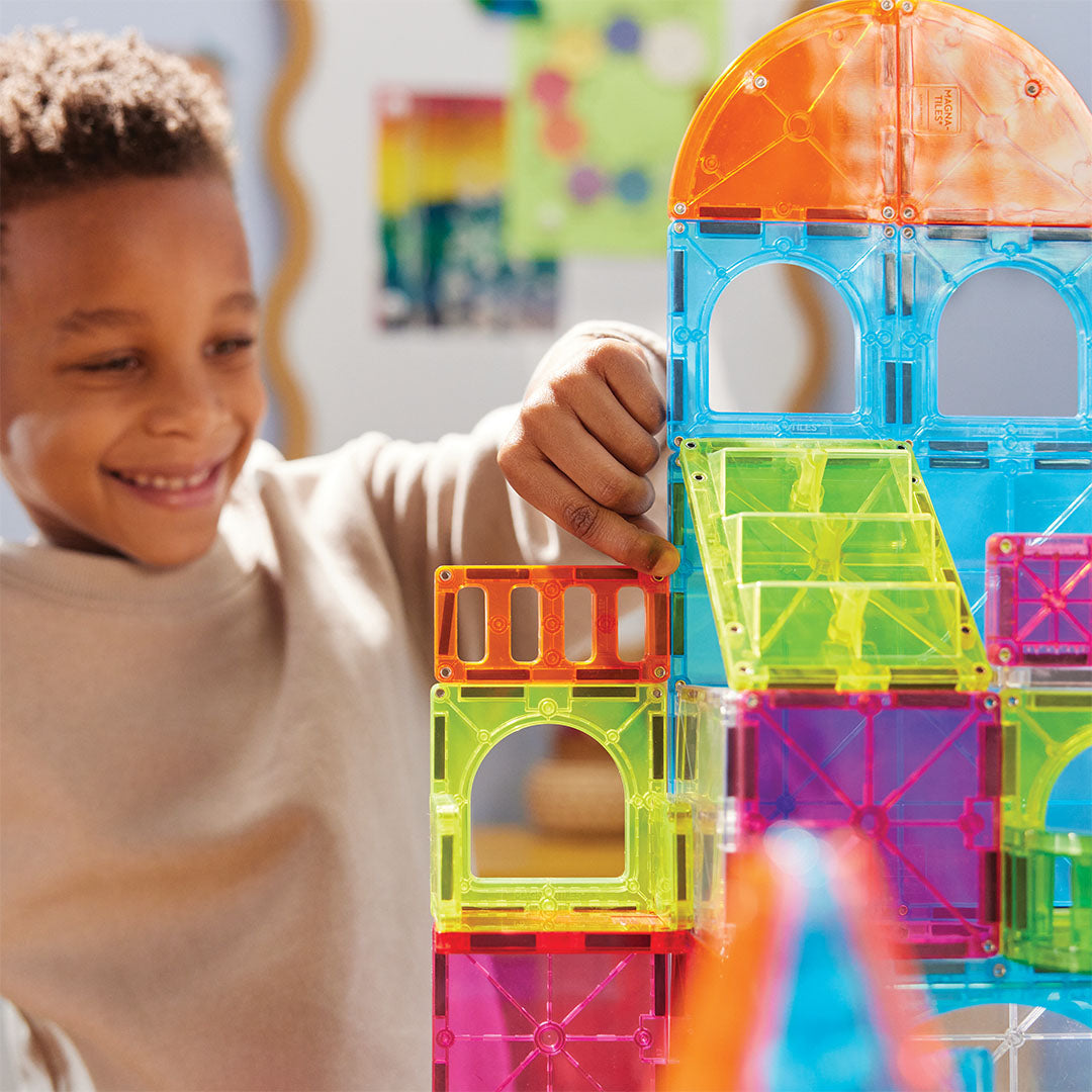 Een lachende jongen bouwt met de Magna-Tiles City Center set en voegt een kleurrijke magnetische tegel toe aan een gebouw.