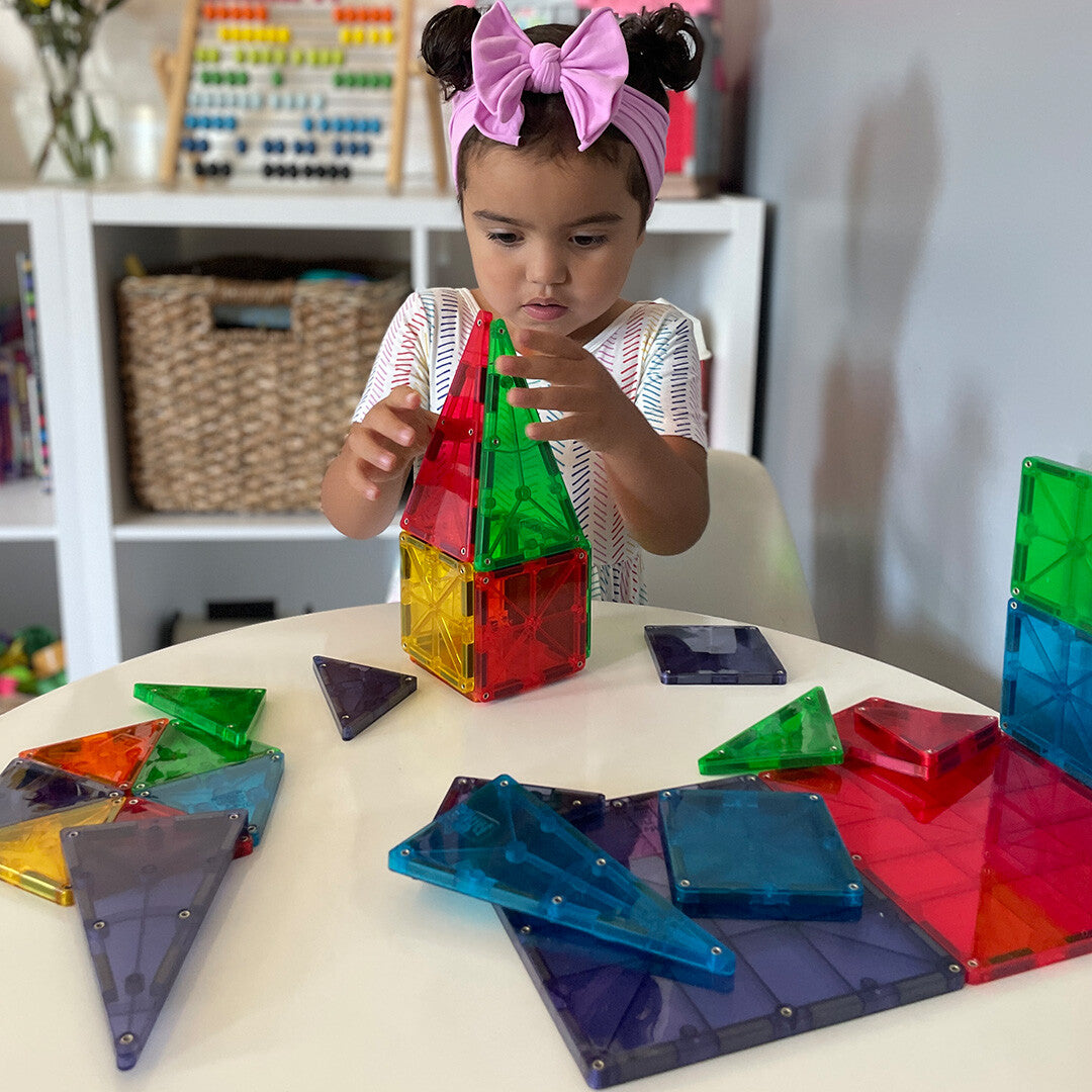 Een meisje bouwt een toren van Magna-Tiles magnetische tegels aan een witte tafel.