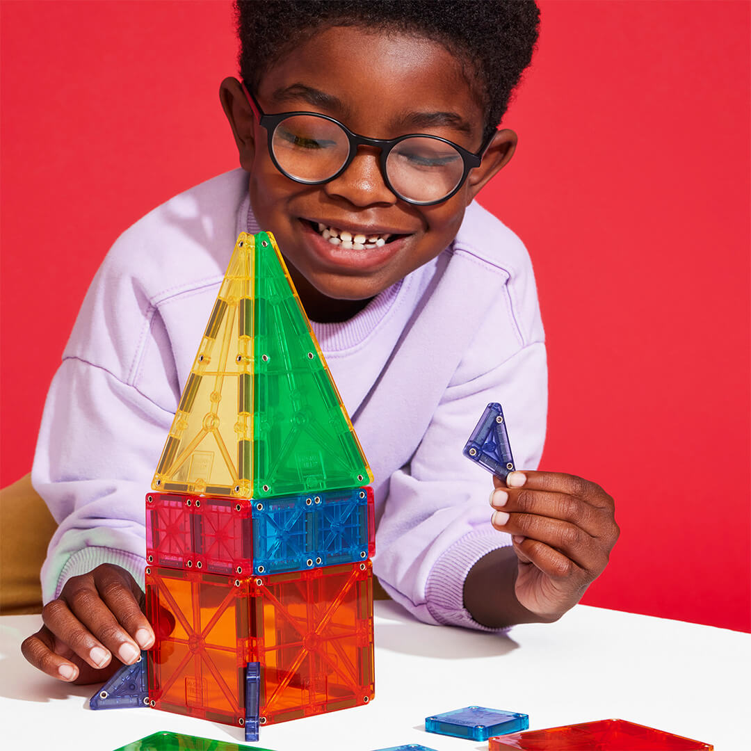 Een lachend kind met bril bouwt een raket van kleurrijke Magna-Tiles magnetische tegels op een witte tafel.