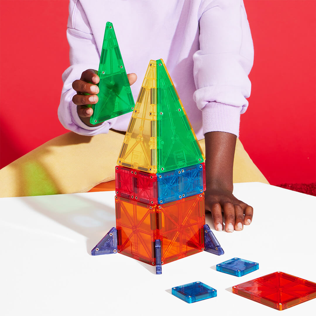Kinderhanden plaatsen een groene Magna-Tiles magnetische tegel op een raketvormig bouwwerk van gekleurde tegels.