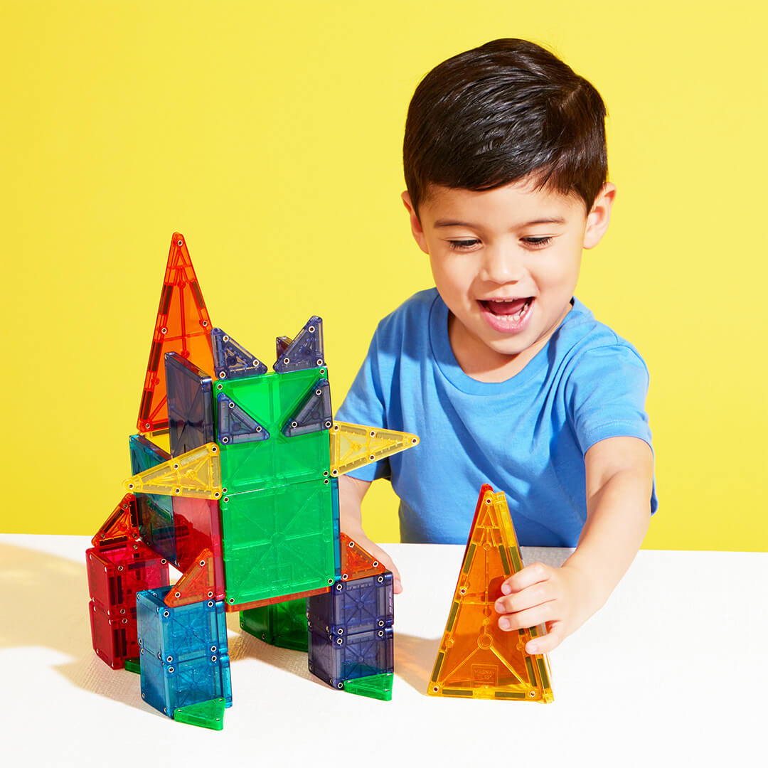 Een lachend jongetje houdt een oranje piramide van Magna-Tiles magnetische tegels vast, naast een fantasiefiguur.