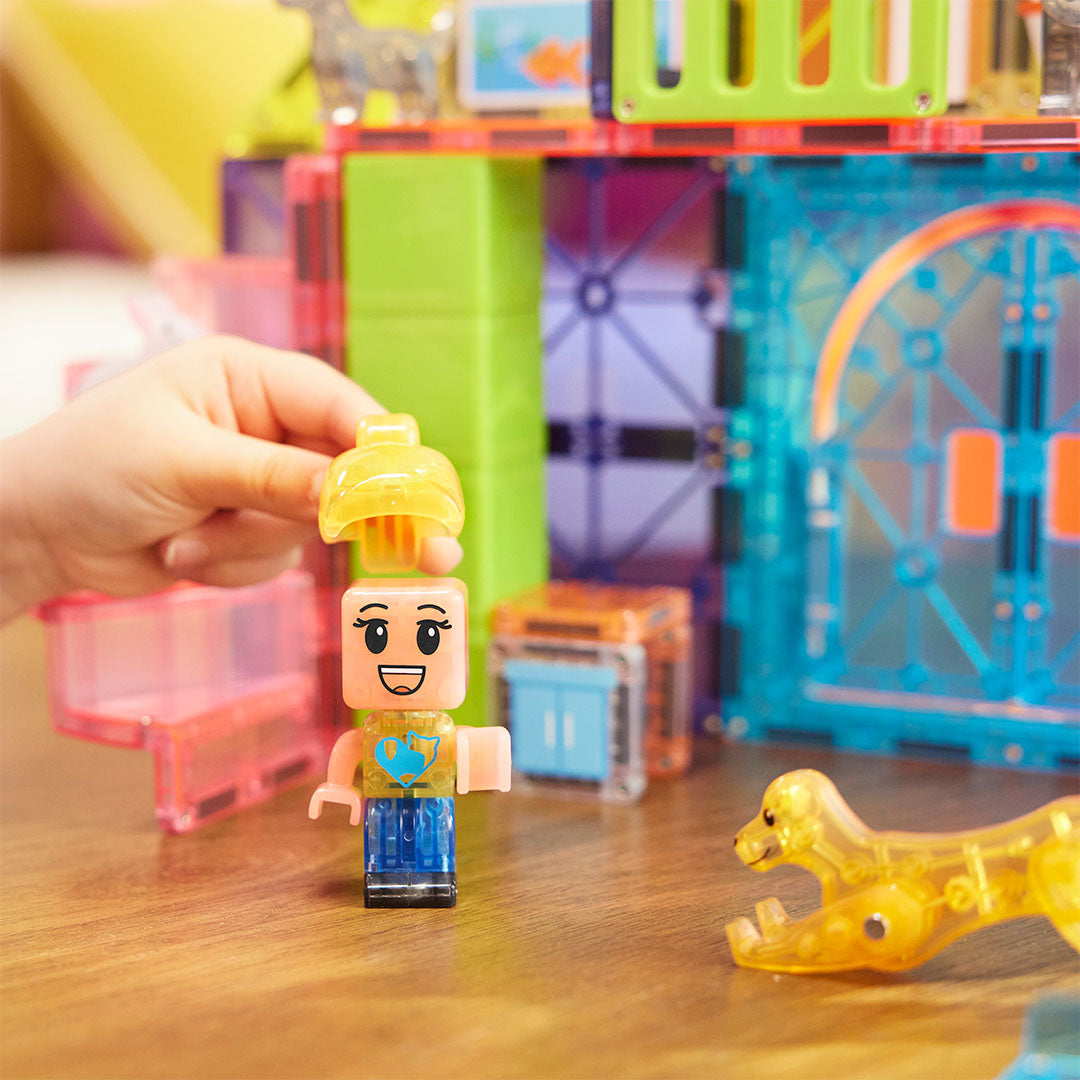 Een kinderhand plaatst een geel haarstukje op een kindfiguur van de Magna-Tiles Pet Playhouse bouwset.