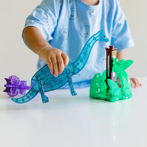 Een kinderhand houdt een blauwe Brachiosaurus dinosaurusfiguur vast uit de Magna-Tiles bouwset.