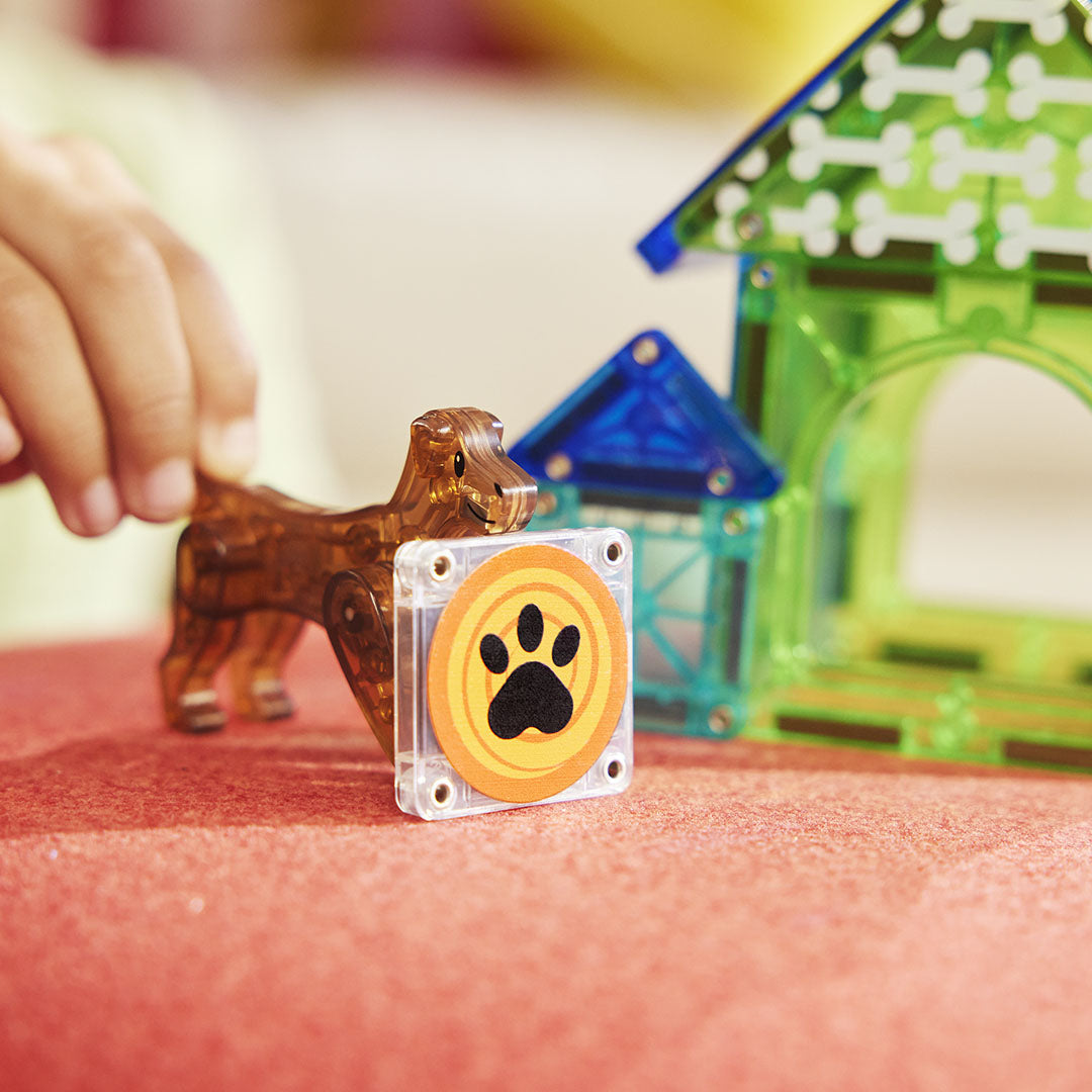 Een kinderhand pakt de bruine hond van de Magna-Tiles Dog House bouwset naast een tegel met pootafdruk.