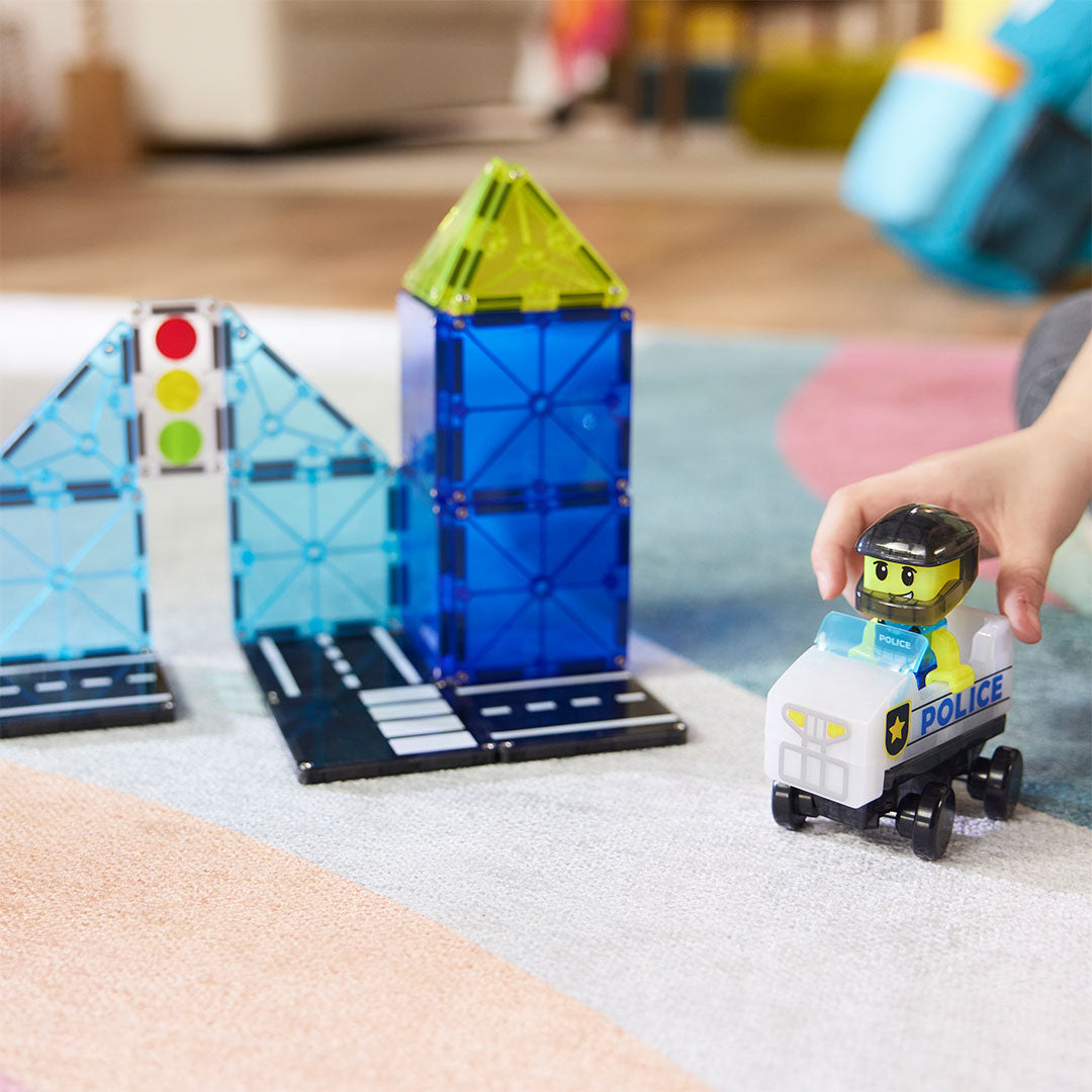 Een kinderhand speelt met de Magna-Tiles politiewagen en agent op een kleurrijk vloerkleed.