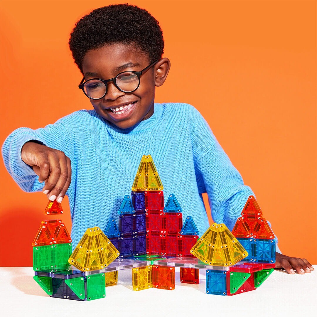 Een lachend kind bouwt een kleurrijk gebouw met Magna-Tiles Micro Mags magnetische tegels.