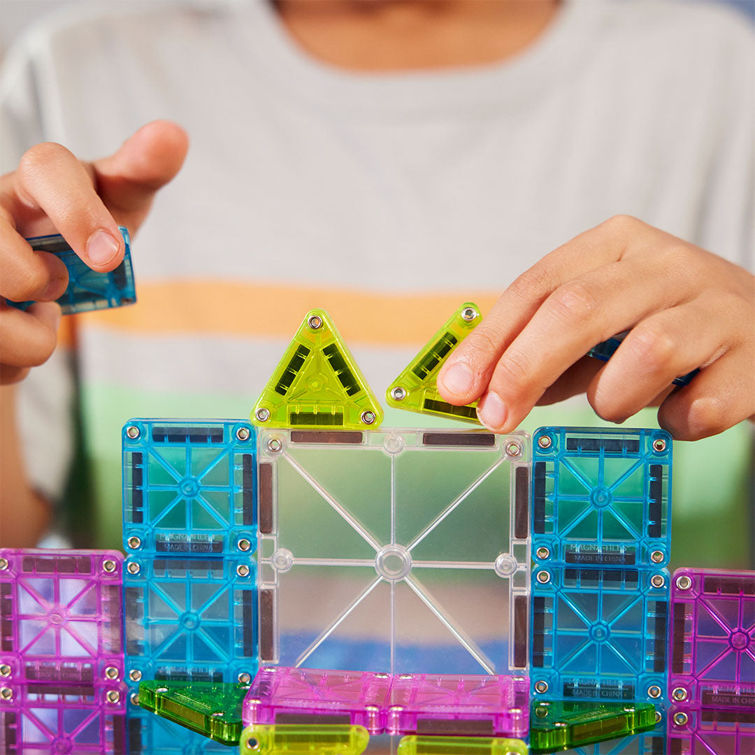 Detail van handen die groene driehoekige Magna-Tiles magnetische tegels aan een kleurrijke constructie bevestigen.