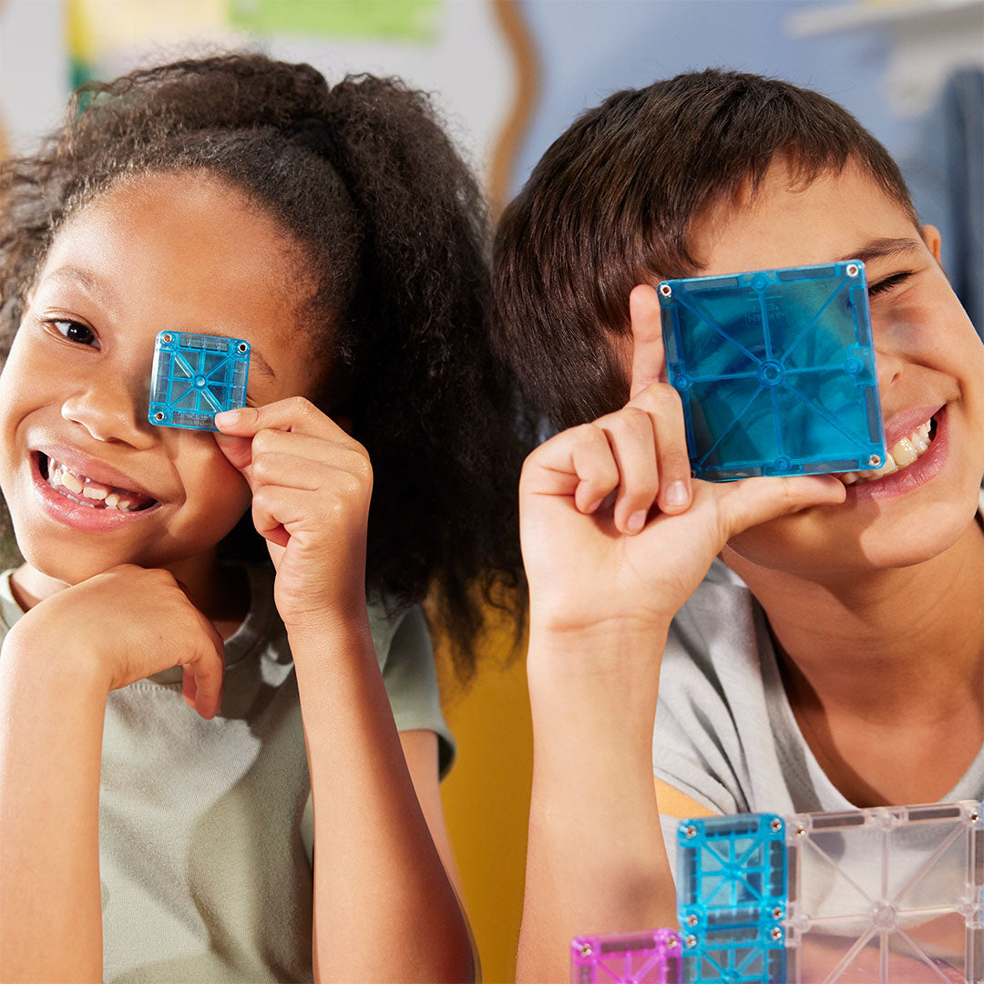 Twee lachende kinderen houden blauwe Magna-Tiles magnetische tegels voor hun ogen.