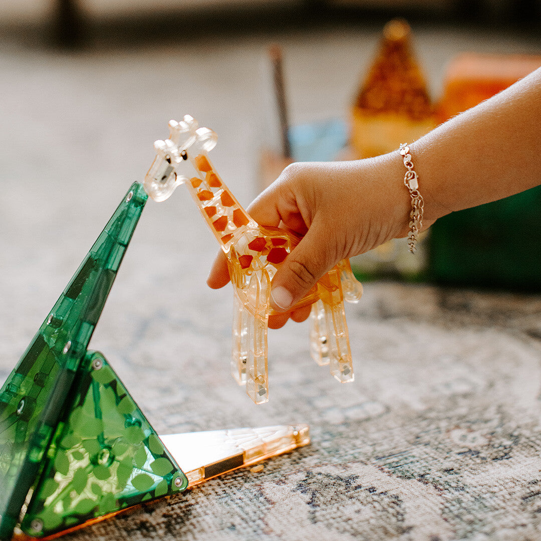 Een kinderhand houdt een doorzichtige, oranje giraf van magnetische tegels vast, naast een groene tegelstructuur.