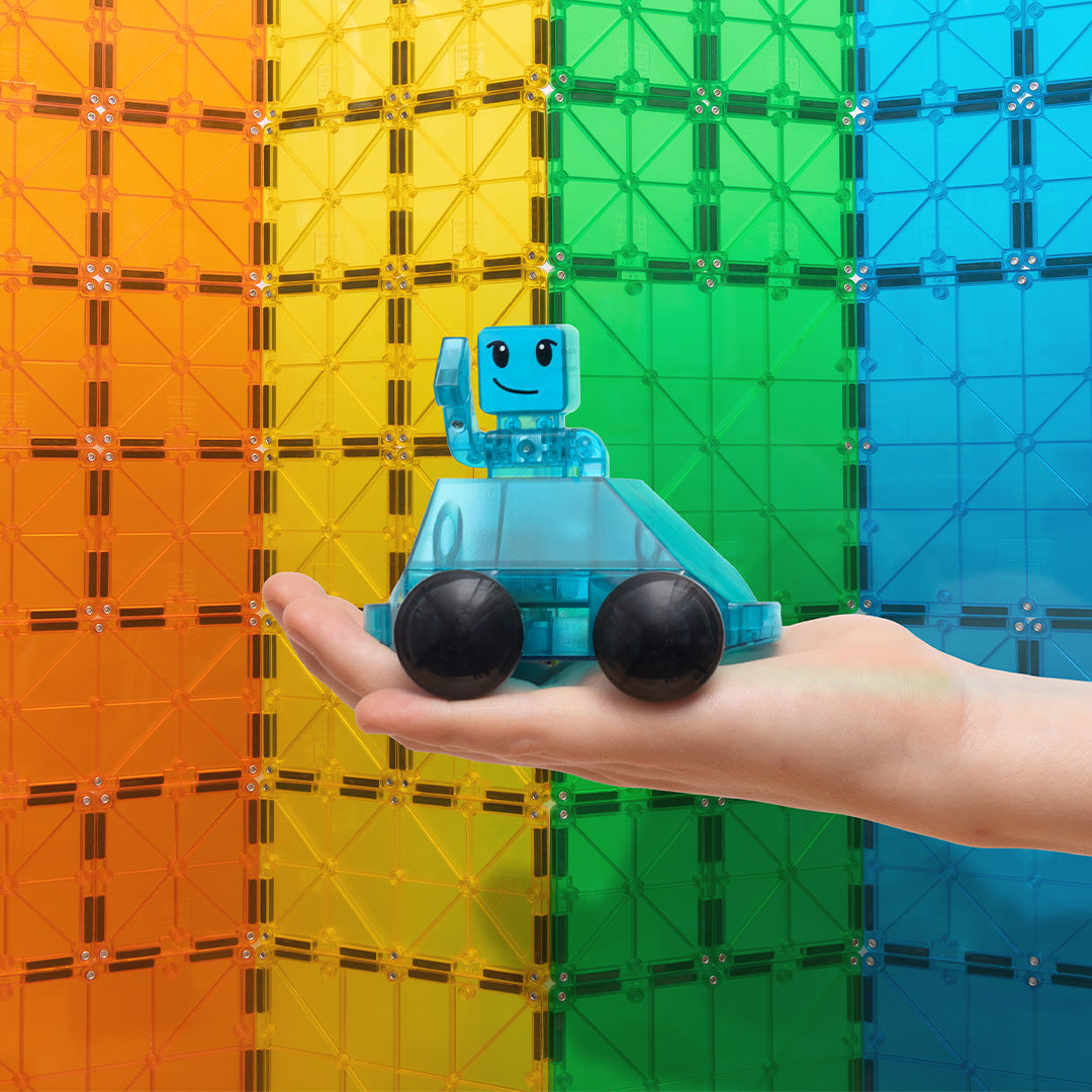 Een hand houdt een blauwe Magna-Tiles Dasher speelgoedauto met robotje vast voor een achtergrond van kleurrijke magnetische tegels.