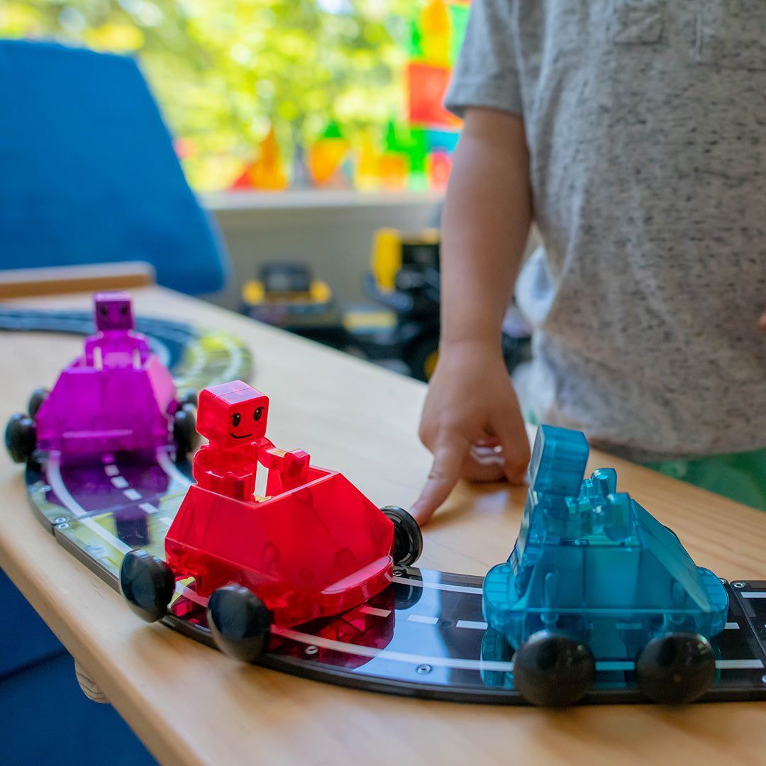 Een kind wijst naar een rode Magna-Tiles Dasher speelgoedauto met robotje op een magnetische weg, met twee andere auto's ernaast.