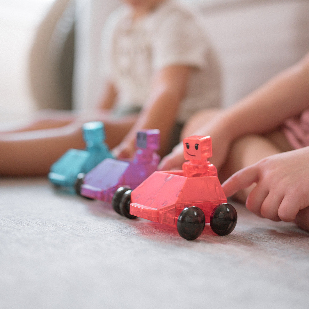 Drie Magna-Tiles Dashers speelgoedauto's met robotjes staan op een rij op de vloer, met spelende kinderen op de achtergrond.